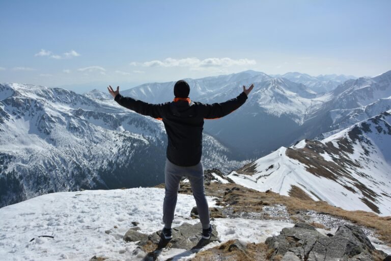 A man stands on a snowy mountain peak with arms raised, embracing the breathtaking view.